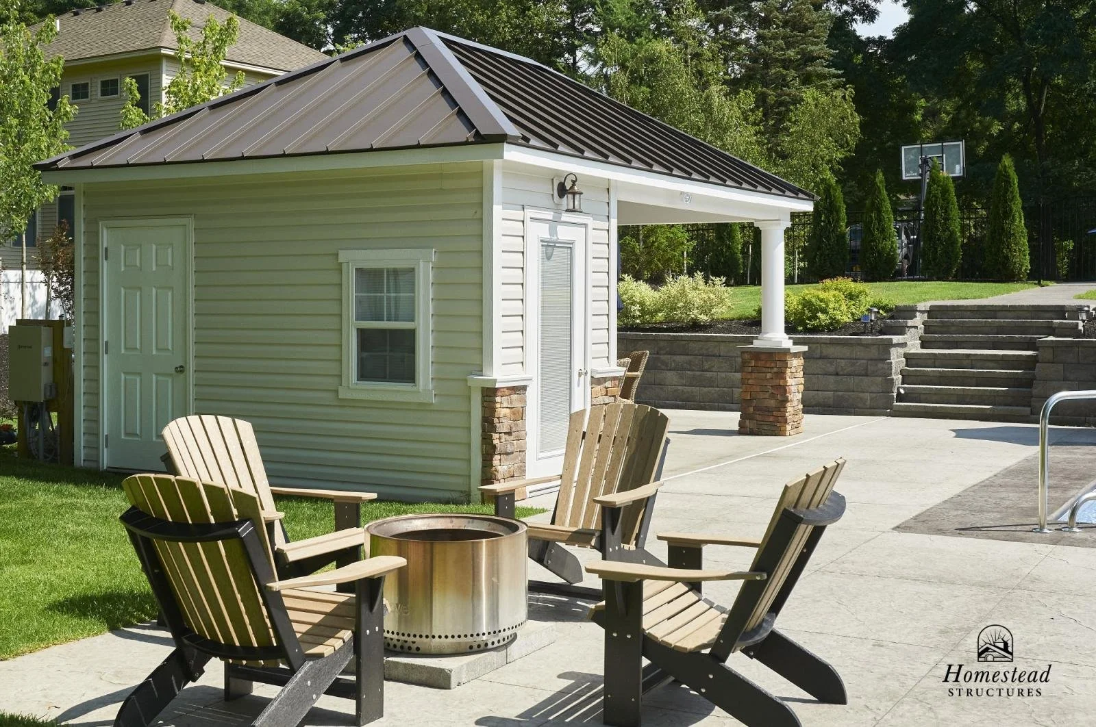 A backyard with a small white building, four Adirondack chairs around a fire pit, and stairs leading up to a garden with bushes and trees.