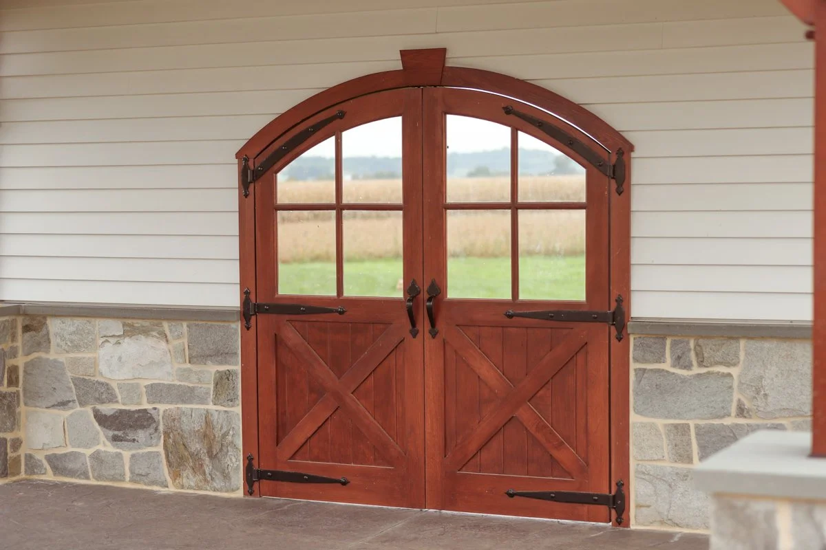 Wooden barn doors with black hardware, set against a wall with stone and siding, opening to a field with grass and a distant treeline.