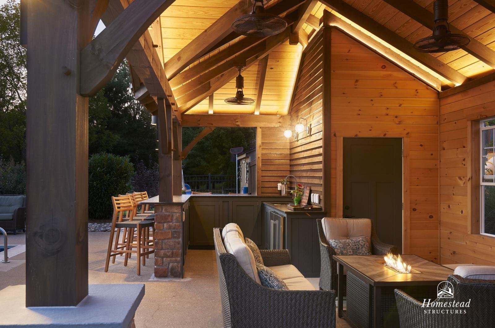 Outdoor patio area with a wooden ceiling, ceiling fans, and cozy seating, including chairs and a table with a fire feature. There is a bar area with stools and a countertop in a backyard setting at dusk.