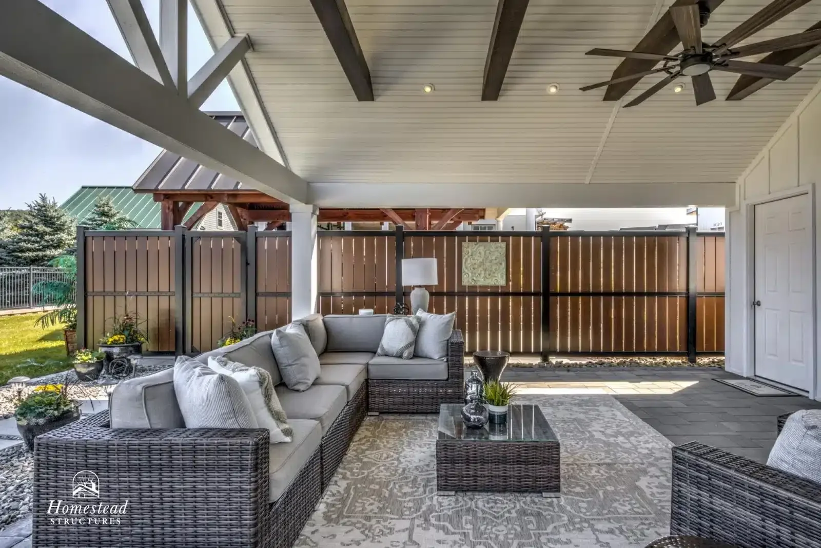 Covered outdoor patio area with beige sectional sofa, wicker chairs, and a glass-top coffee table on a patterned rug, surrounded by a wooden fence, with ceiling fan and some potted plants visible.