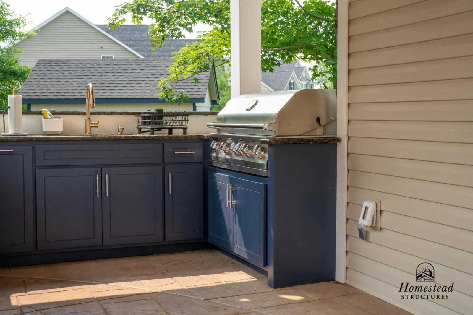 Outdoor kitchen with a stainless steel grill, dark blue cabinetry, a granite countertop, a sink, dish drying rack, and a digital thermostat on the wall. There are trees and rooftops of neighboring houses in the background.