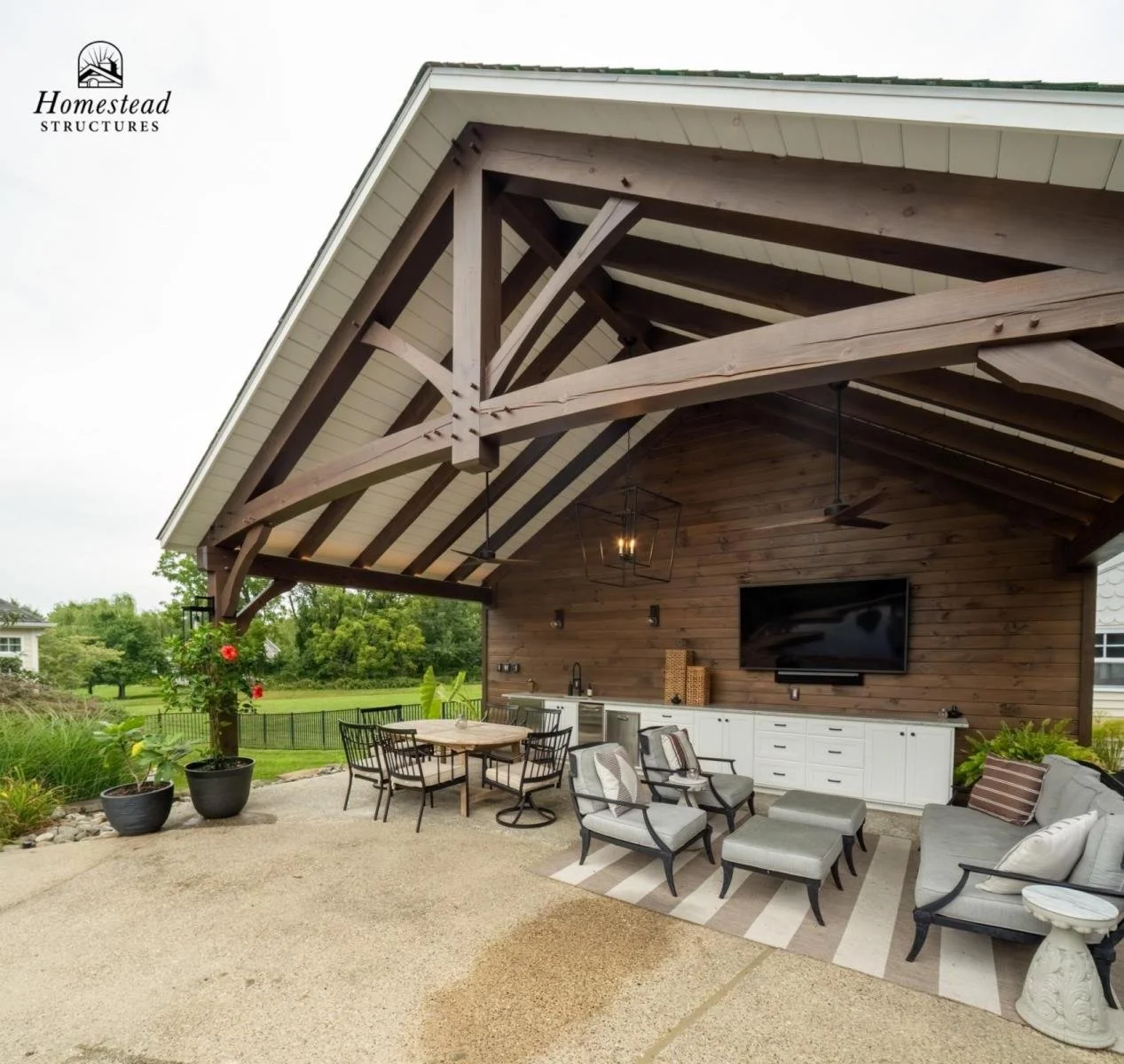 Outdoor covered patio with seating, dining table, TV, and decorative plants, featuring a wooden ceiling and natural greenery in the background.