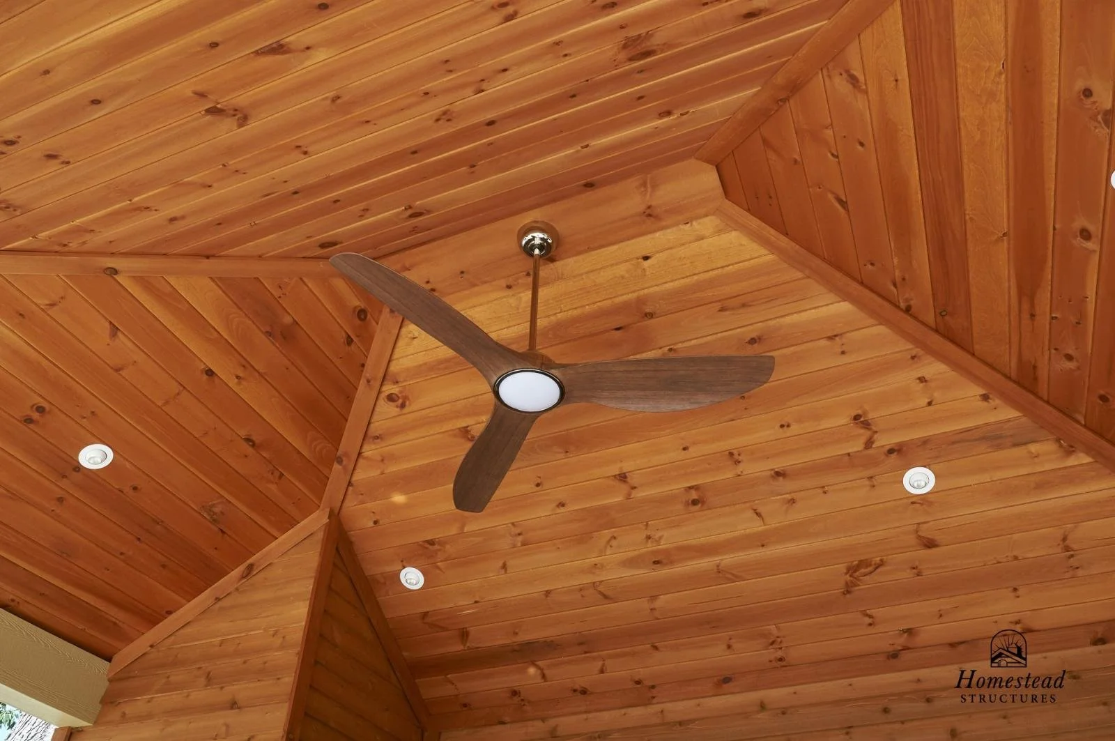 Wood-paneled ceiling with a modern ceiling fan featuring three dark wooden blades and a central light fixture, along with three recessed lights and a logo that says "Homestead Structures."