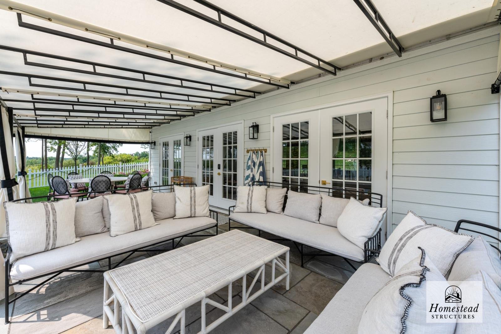 Outdoor covered patio with white siding, black metal furniture with white cushions, and a view of green trees and a white picket fence in the background.