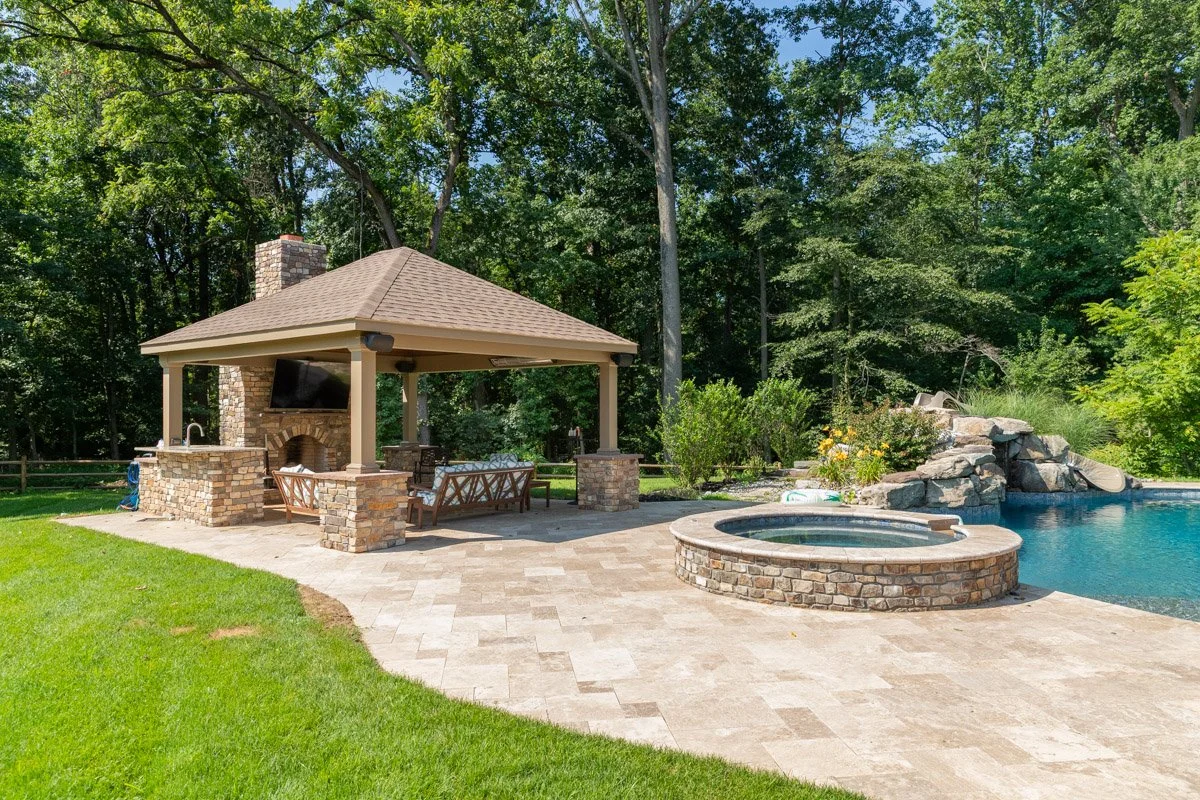 Backyard with swimming pool, hot tub, stone waterfall, and shaded outdoor living area surrounded by green trees.