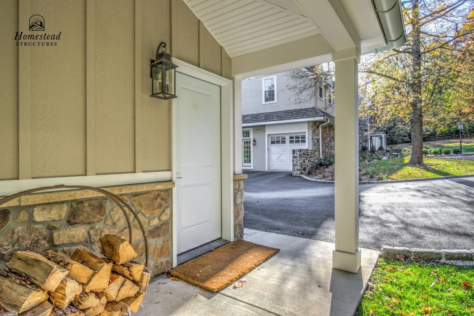 View of a front porch with a white door, a black outdoor lantern light, a piled stack of firewood, and a brown doormat. The porch is part of a house with beige siding and stone accents. In the background, there is a curved driveway, a garage, a large