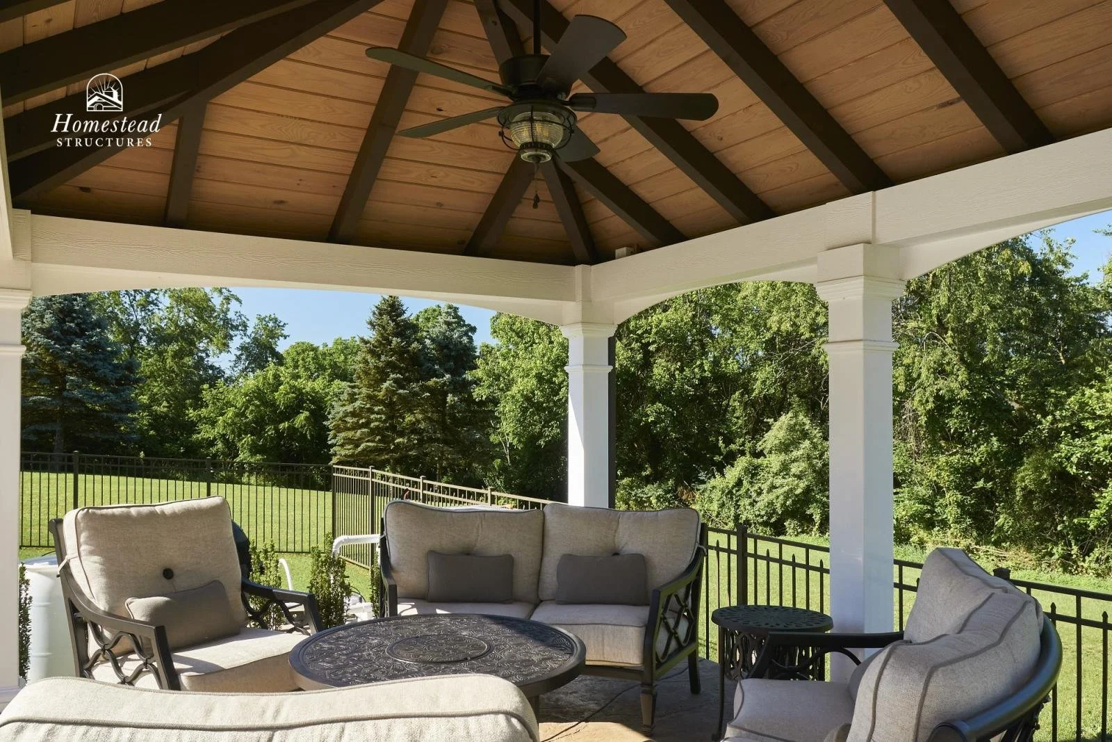 Covered porch area with outdoor furniture, including cushioned chairs and a round table, overlooking a lush green yard with trees and a black metal railing.