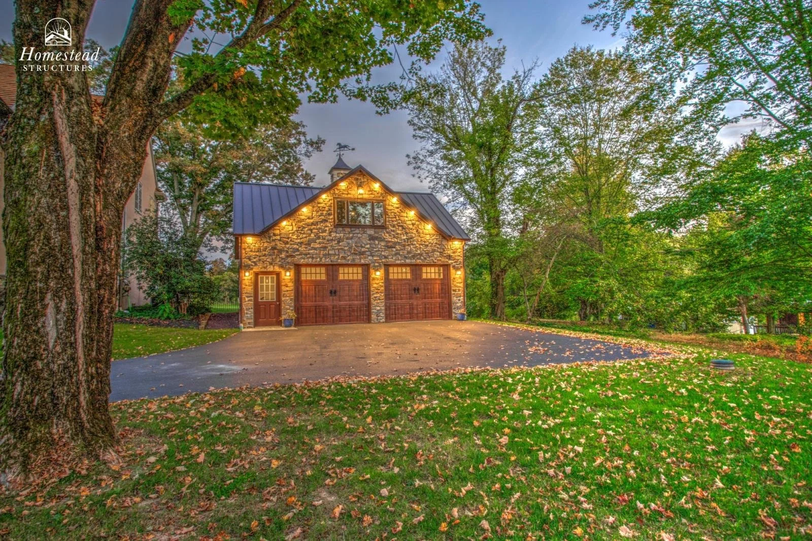 A stone garage with wooden doors illuminated by outdoor lighting, surrounded by trees with green leaves and a lawn with fallen leaves during dusk.