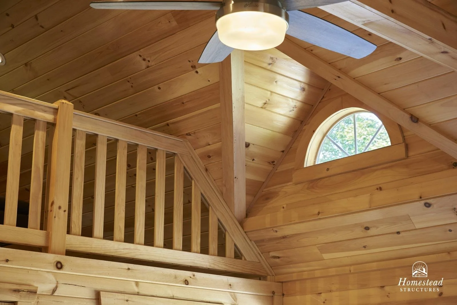 Interior view of a wooden cabin with a sloped roof, a small round window, a ceiling fan with a light, and a wooden railing on a loft or upper level.