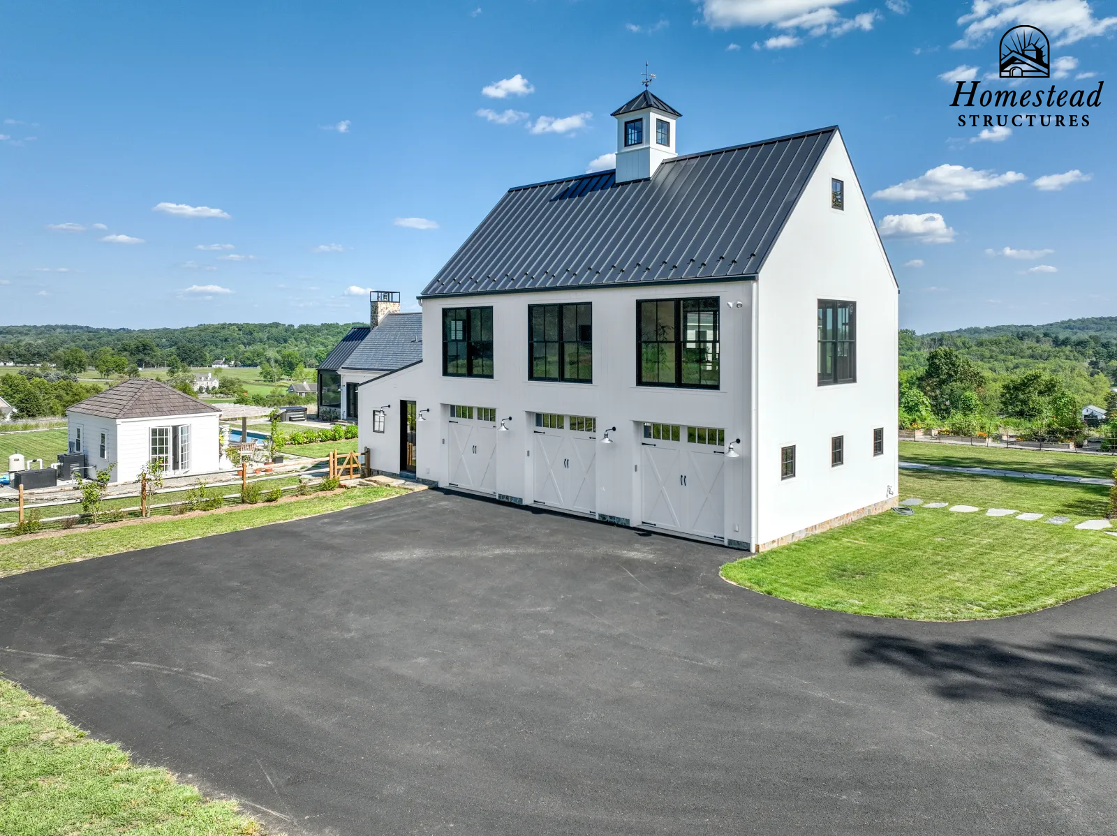 Modern white barn with black roof and large windows, surrounded by a green landscape and a clear blue sky, with a small outbuilding nearby.