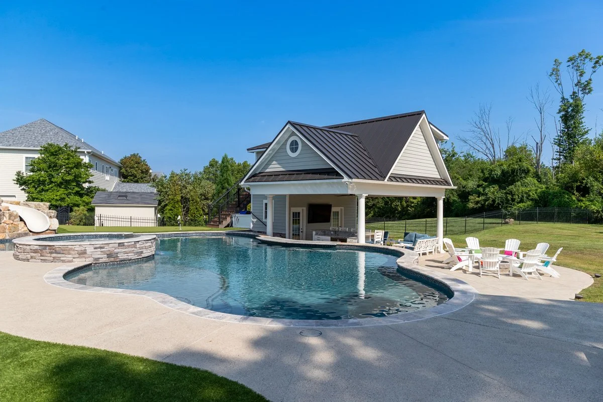 A backyard with a freeform swimming pool, a pool house with a steep roof, seating area with white Adirondack chairs, and a waterslide on the left, surrounded by green grass and trees under a clear blue sky.