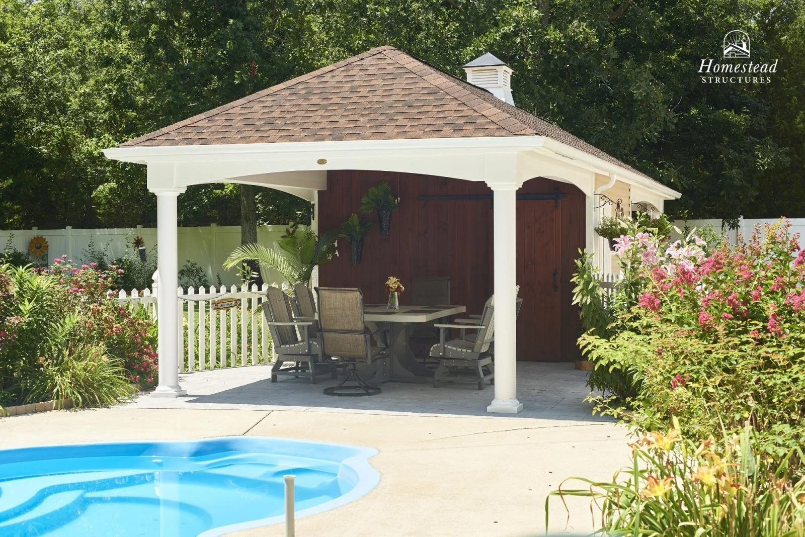 A backyard patio with a white gazebo, outdoor table and chairs, lush flower landscaping, swimming pool in the foreground, and a wooden shed with a red door in the background. There is a white fence surrounding the yard, and dense trees beyond.