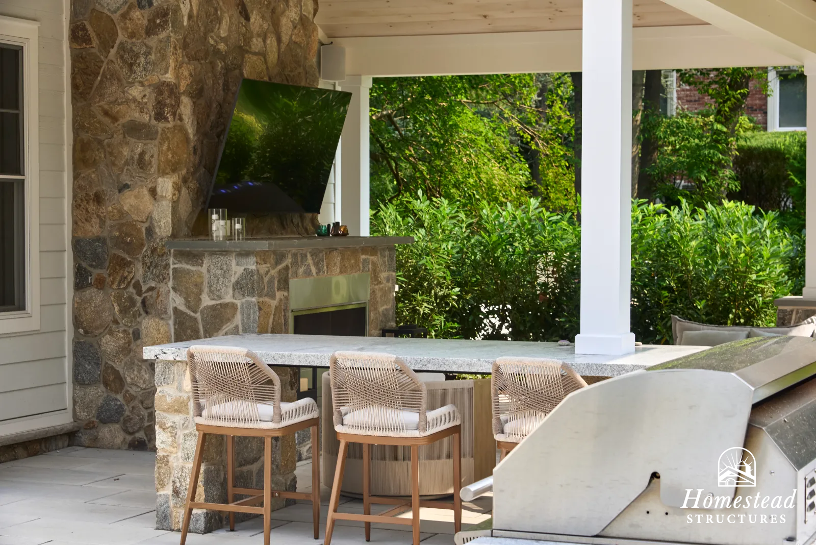 Outdoor patio with a stone fireplace, mounted flat-screen TV, a granite countertop bar with wicker chairs, surrounded by green bushes and trees.