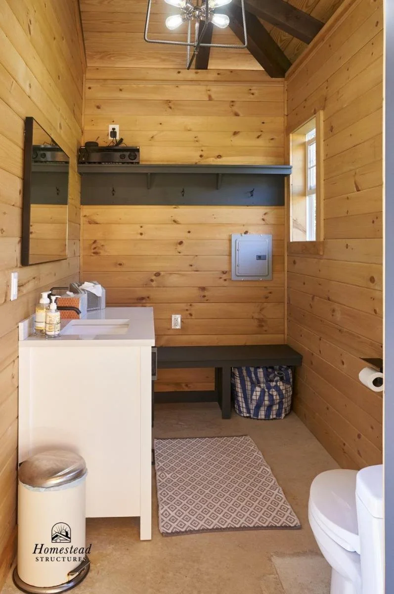 Small bathroom with wood-paneled walls, a window, a mirror, a small sink area with soap and sanitizer bottles, a trash can, a bench with a striped bag underneath, a gray shelf, a small electrical panel on the wall, a toilet, and a patterned rug on th