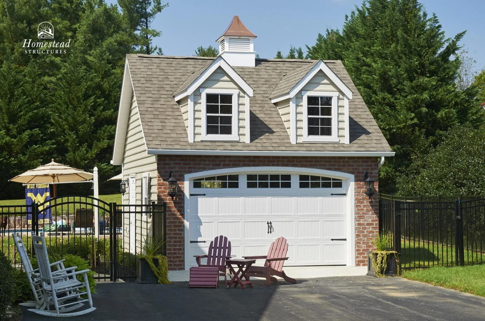 A small two-story garage with a brick lower wall and beige siding upper wall, featuring two dormer windows on the roof. In front, there's a driveway with two pink Adirondack chairs and a small matching table. A black metal gate and fence surround the
