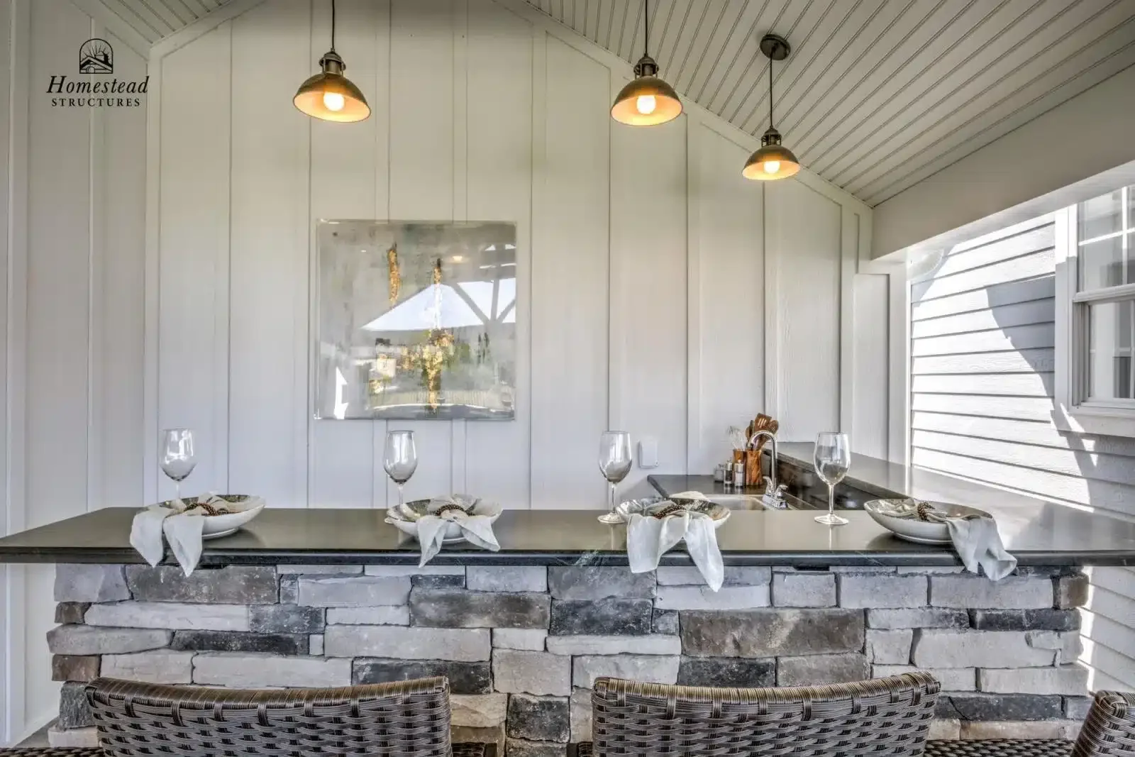 A kitchen counter with four wine glasses, each placed on a plate with a white napkin and cutlery, and a stone facade base, in front of white paneled walls and hanging pendant lights.