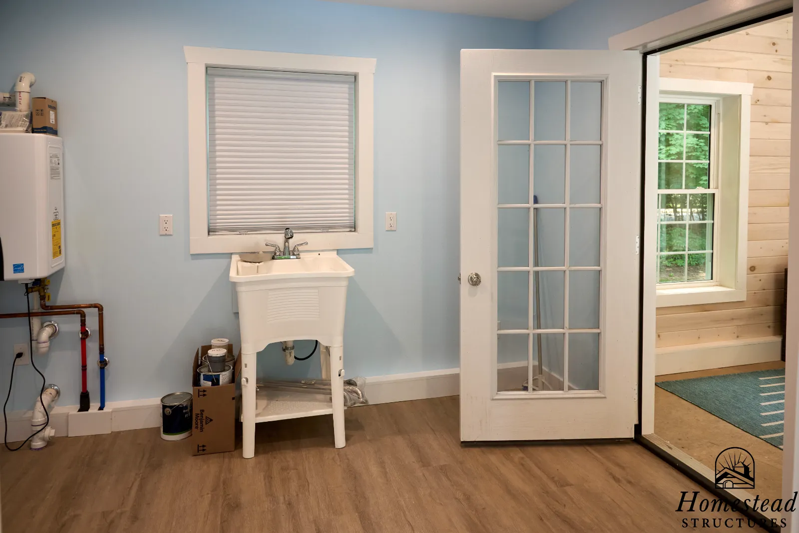 Pool house interior view of a laundry room with a utility sink, a window with closed blinds, a door with glass panes leading to the outside, a water heater on the wall, and wood flooring.