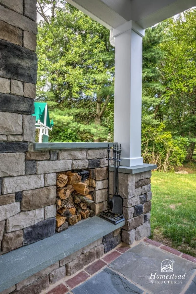 A stone porch with a built-in wood storage area filled with chopped firewood. There are fireplace tools hanging on the wall next to the wood storage and a partial view of a house with green roofs in the background. The setting is outdoors with trees 