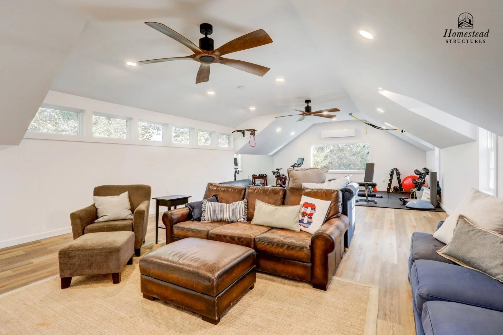Living room with brown leather sofa, blue and beige armchairs, wooden flooring, ceiling fans, and workout equipment in the background.