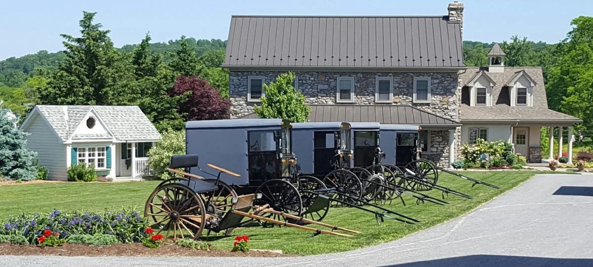 Four horse-drawn carriages parked on a lawn in front of a stone and wood house, with a smaller white building nearby, surrounded by trees and flowers.