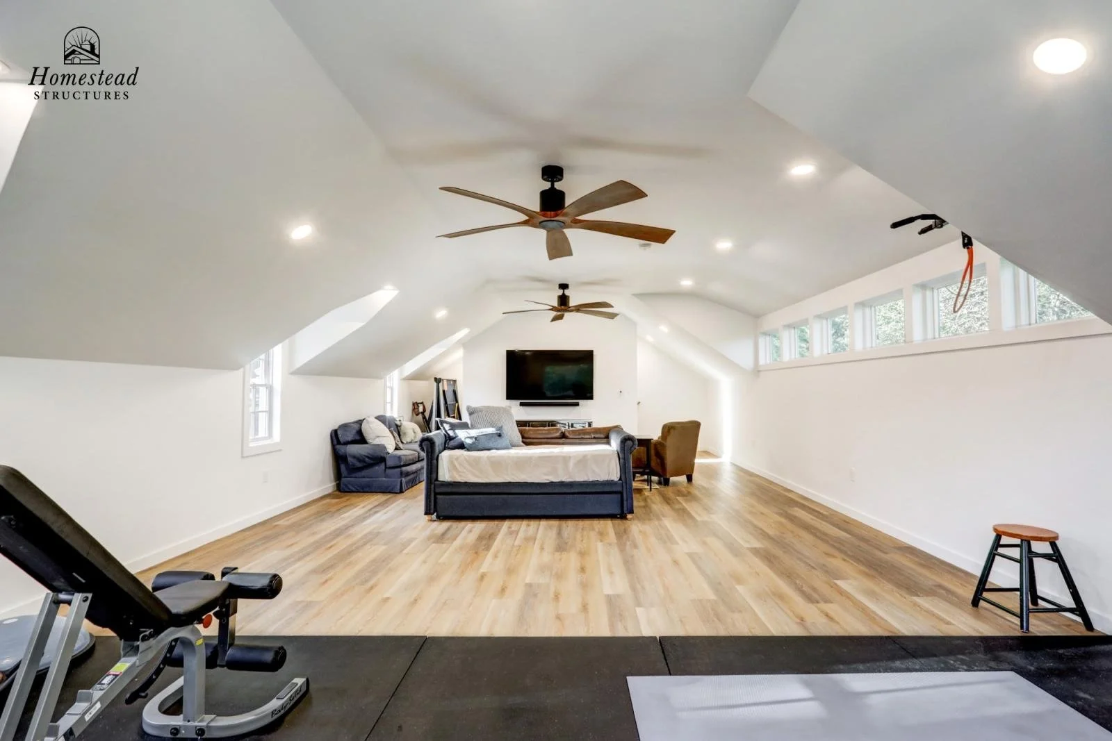 Interior of a spacious, well-lit attic living room with hardwood flooring, white walls, ceiling fans, a wall-mounted TV, sofas, chairs, and windows near the ceiling.