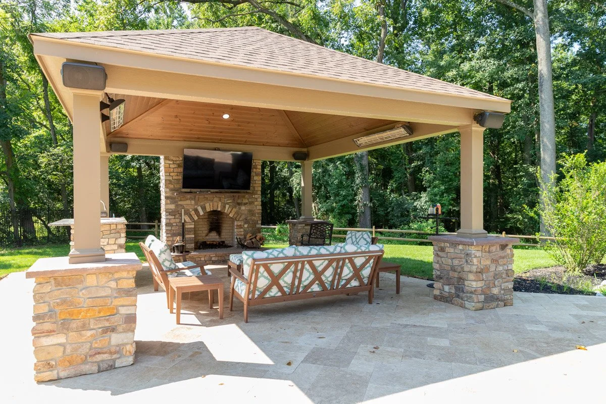 Outdoor pavilion with a stone fireplace, a mounted TV, and patio furniture, surrounded by trees and a grassy yard.