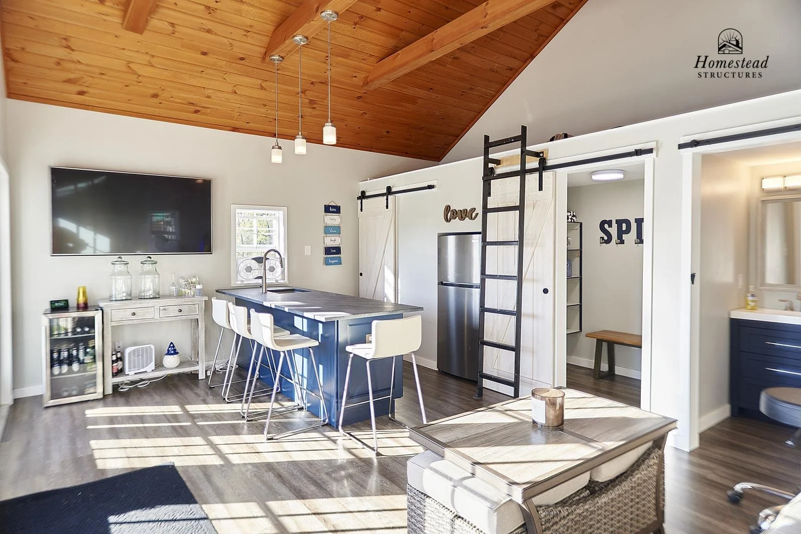 Open-concept kitchen and living space with a vaulted wooden ceiling, large flat-screen TV, and a kitchen island with four white chairs. There is a stainless steel refrigerator, a sliding barn door, and wall decor including a wooden 'love' sign and a 
