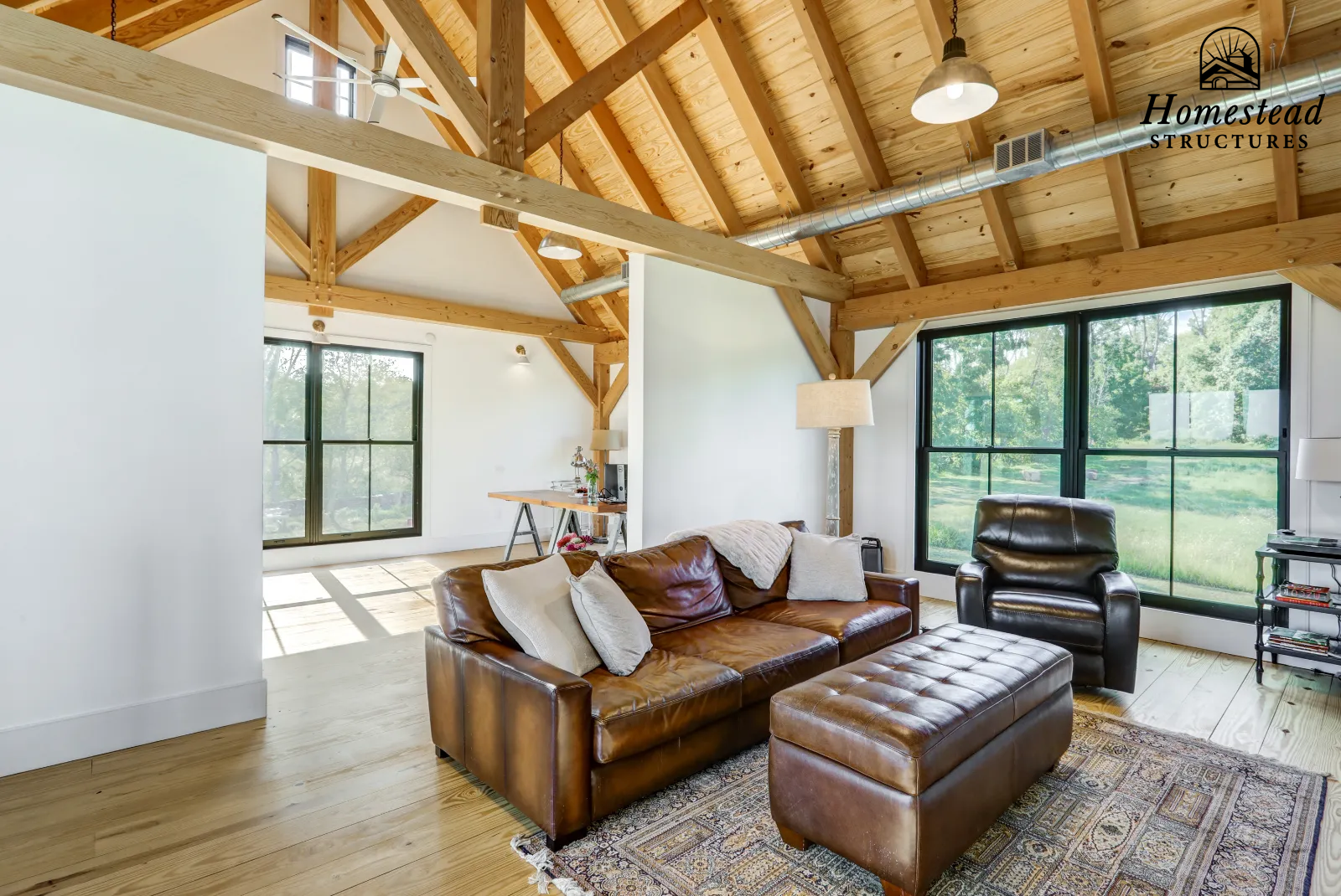 Living room with large windows, exposed wooden beams and ceiling, leather sofa with cushions, armchair, and an ottoman, with a rug on wooden floors.
