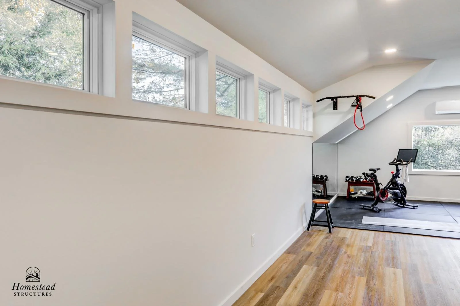 Home gym in a room with five upper windows, a wall mirror, exercise bikes, weights, an exercise mat, and a workout pull-up bar, with wood flooring and a small stool.