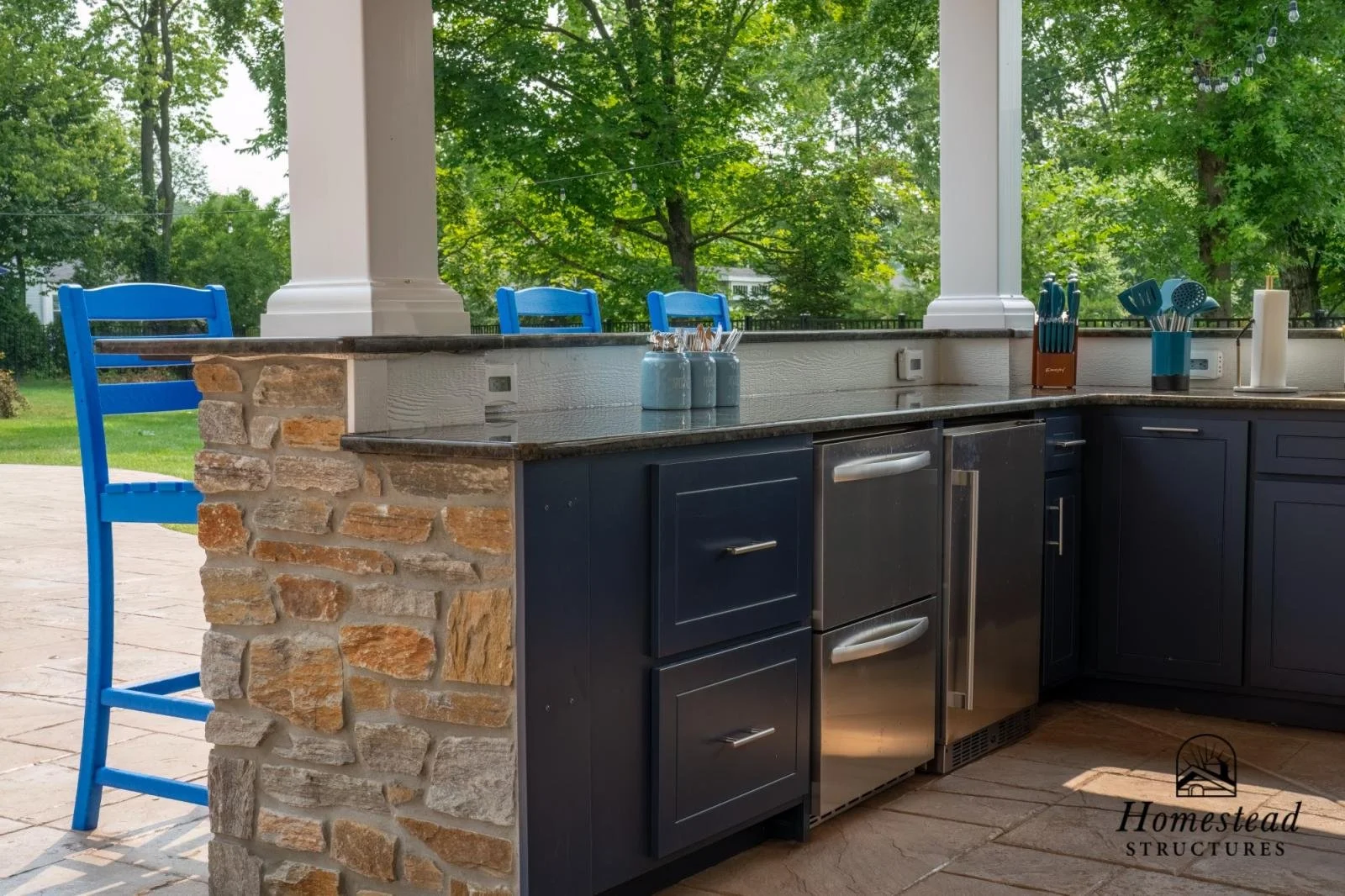 Outdoor kitchen with a stone and wood counter, blue chairs, and greenery in the background.