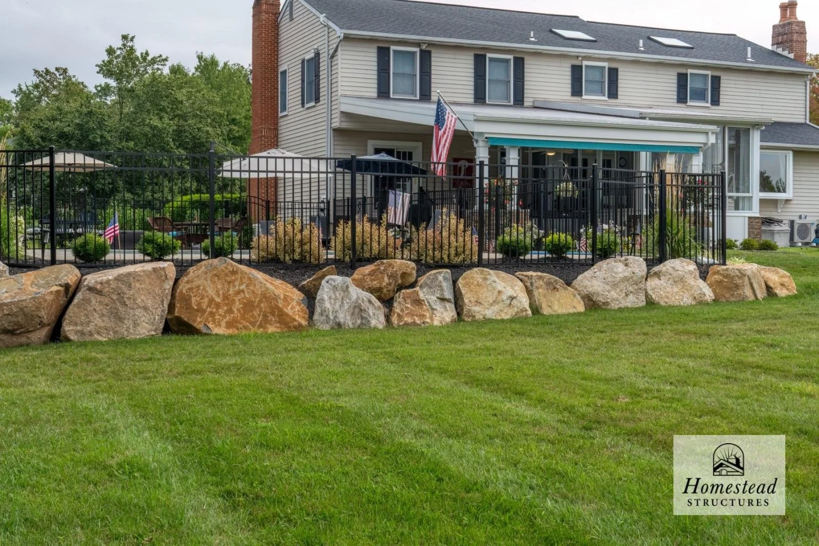 Backyard patio area enclosed by black metal fence, with large rocks along the edge, lush green grass in the foreground, and a two-story house with a porch, outdoor furniture, and patriotic decorations in the background.