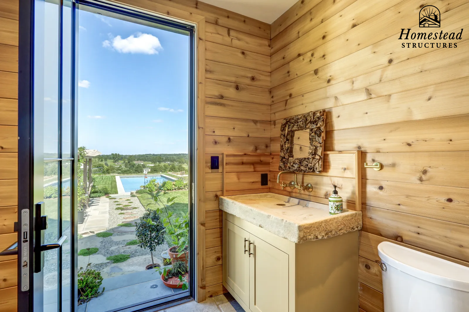 Interior of a rustic bathroom with wooden panel walls, a stone sink vanity, a mirror with a rock frame, a toilet, and an open door showing a backyard with a pool and garden.