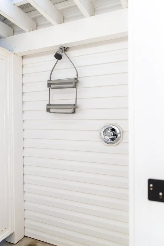 Empty outdoor shower with a showerhead, two-tier soap shelf, and a control knob, all mounted on white horizontal siding.