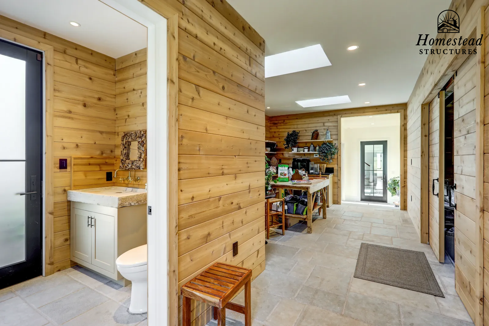 Interior of a wooden house with natural wood paneling on walls, a bathroom with a small sink and a toilet on the left, a hallway with a small wooden stool, and a room with a door at the back, natural lighting from skylights, and shelves with decorati