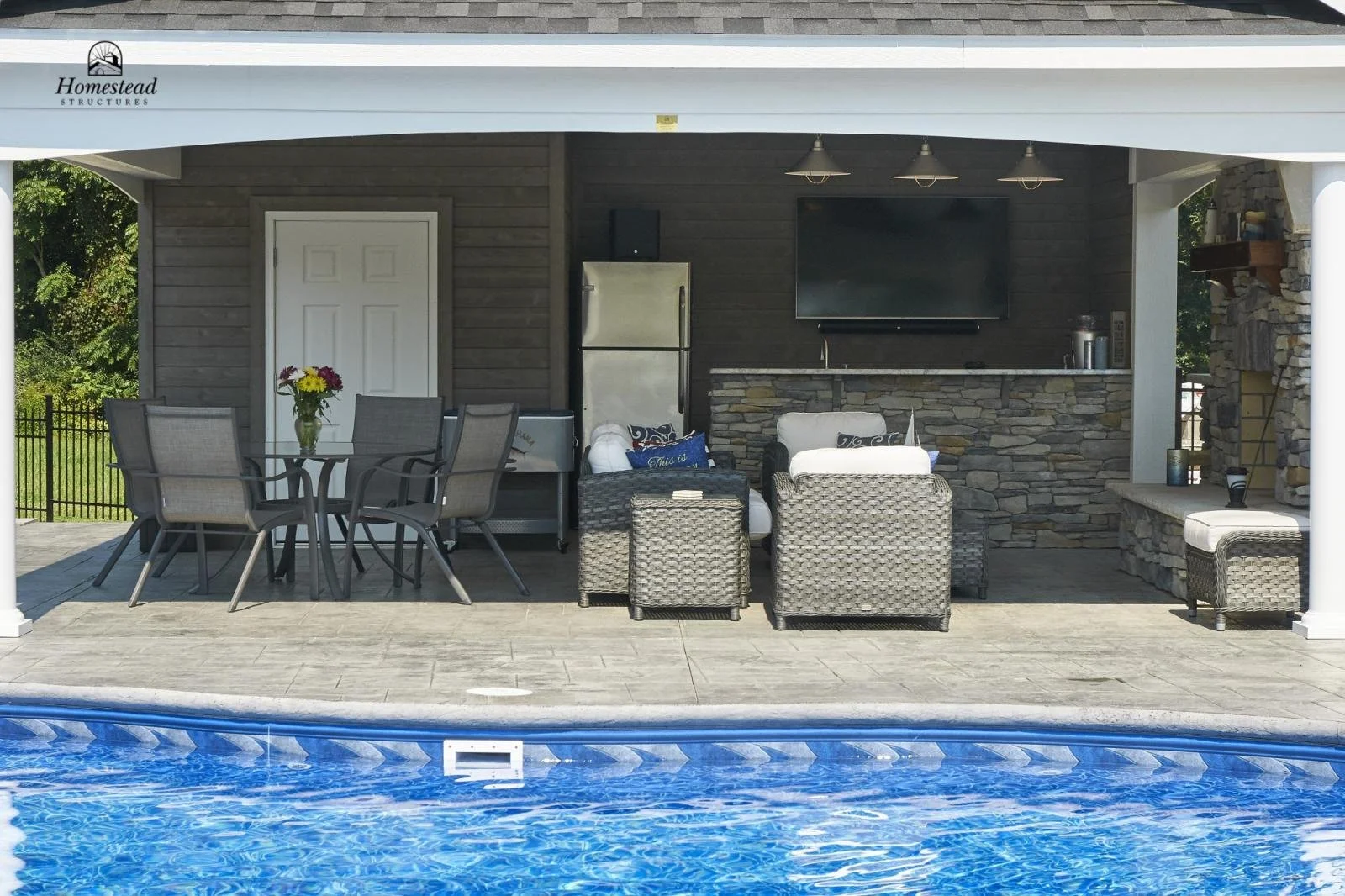 Outdoor patio area with a swimming pool in the foreground, a table with chairs and a flower arrangement, a couch, chairs, a refrigerator, and a large TV mounted on a stone and wood wall in the background.