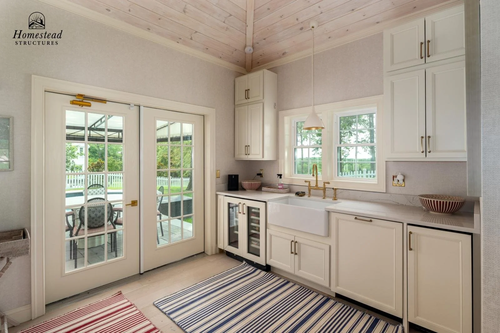 Kitchen with white cabinets, gold fixtures, a farmhouse sink, and a window above it. French doors lead outside to a patio with seating. Two striped rugs are on the light-colored floor, and a wooden ceiling adds warmth.