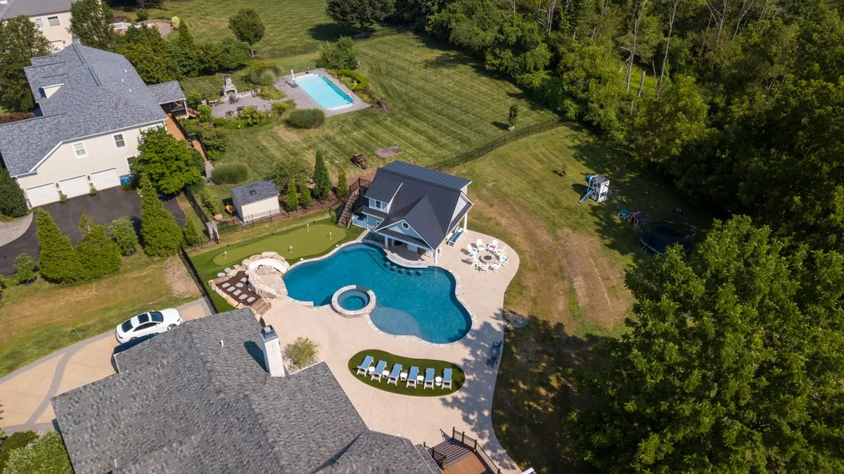 Aerial view of a suburban backyard with a large in-ground swimming pool with a hot tub, a pool house, a patio with outdoor furniture, a children's playset, a lawn, and neighboring houses with a swimming pool and trees.