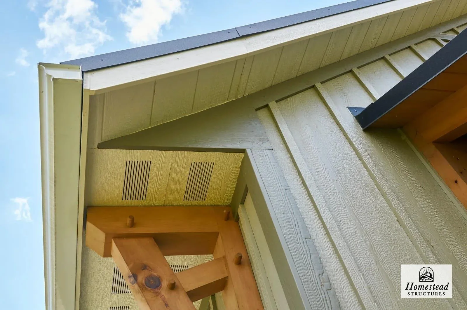 Close-up view of a wooden house exterior with light green painted siding, a black trim, and a wooden beam. The image shows a section of the roof and wall with vents, a corner detail, and a logo for Homestead Structures.