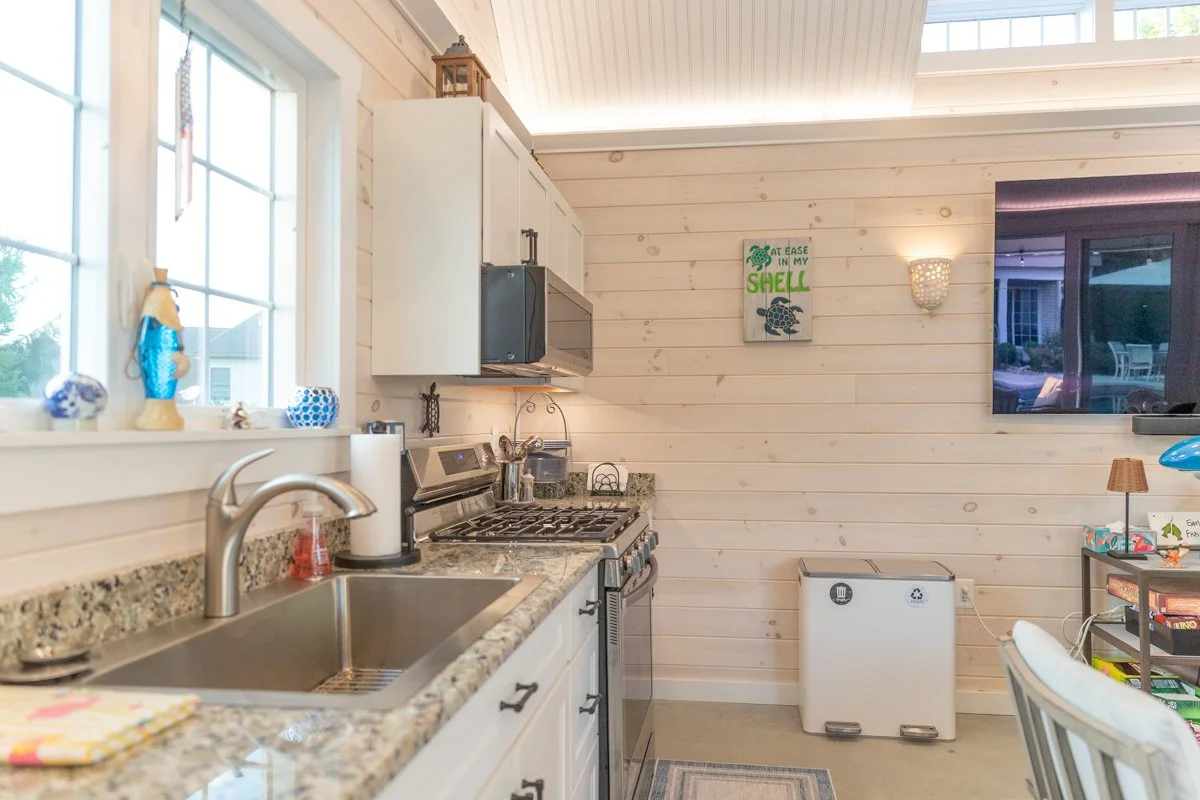A cozy kitchen with white wood-paneled walls, granite countertops, a stainless steel stove, microwave, and sink. Decor includes seashell-themed items and a green turtle art on the wall. A window above the sink shows a residential neighborhood outside