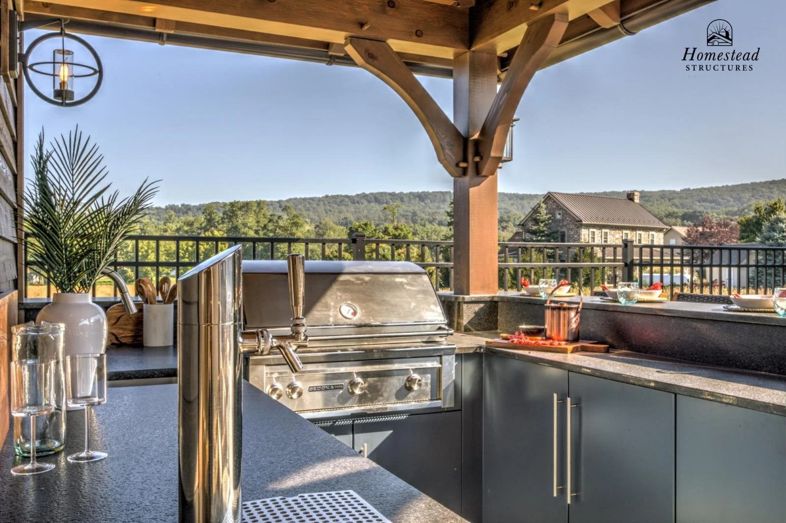 Outdoor kitchen on a deck with a view of rolling hills, featuring a stainless steel grill, counter space, a potted plant, glasses, and a chandelier.