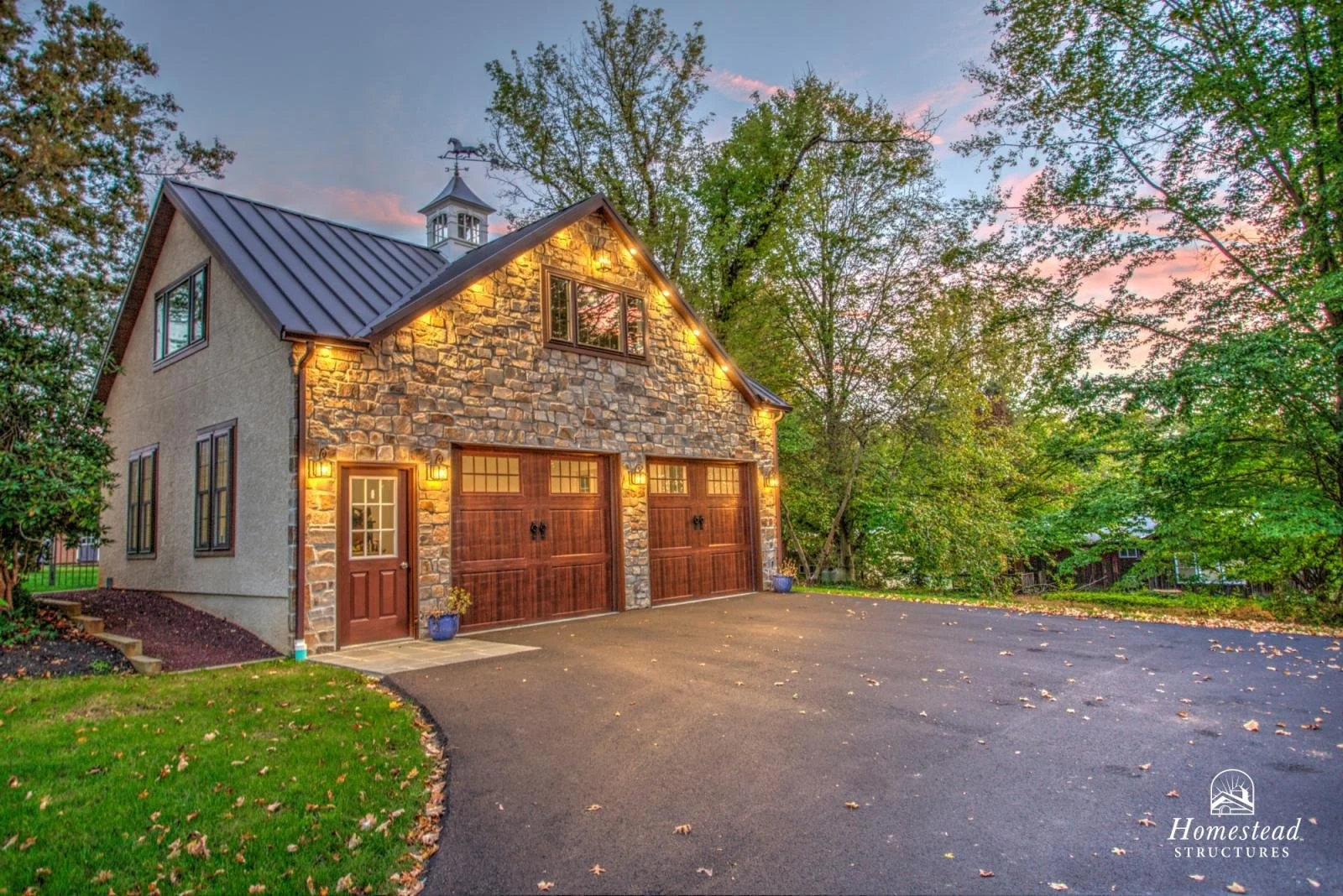 A two-story house with a stone and stucco exterior, illuminated by exterior lights, featuring a double garage with wooden doors, surrounded by trees and a well-maintained lawn, during twilight.