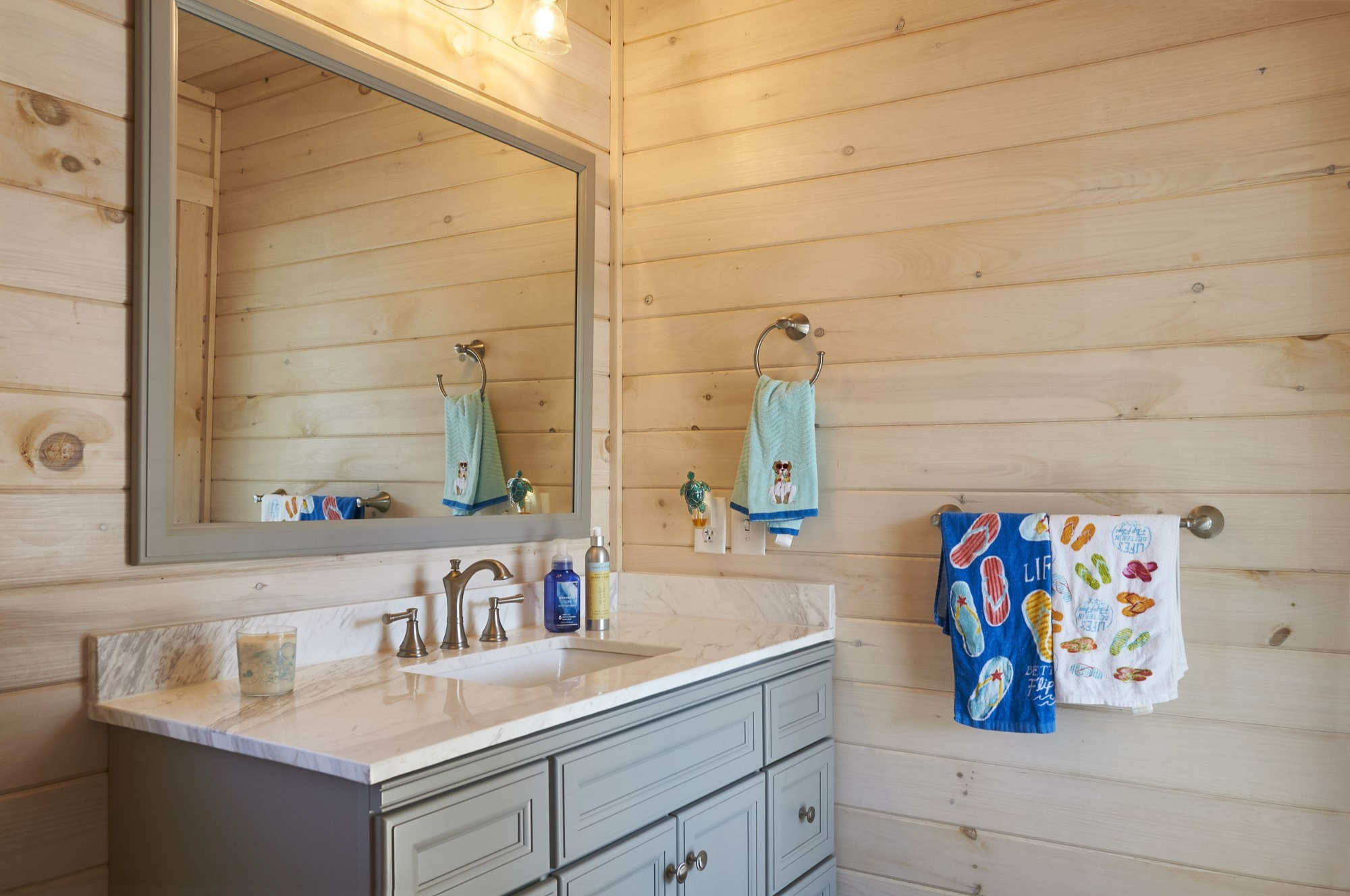 Bathroom with wooden plank walls, large mirror, marble countertop sink, silver faucet, blue and white towels with flip-flop patterns hanging on the wall.