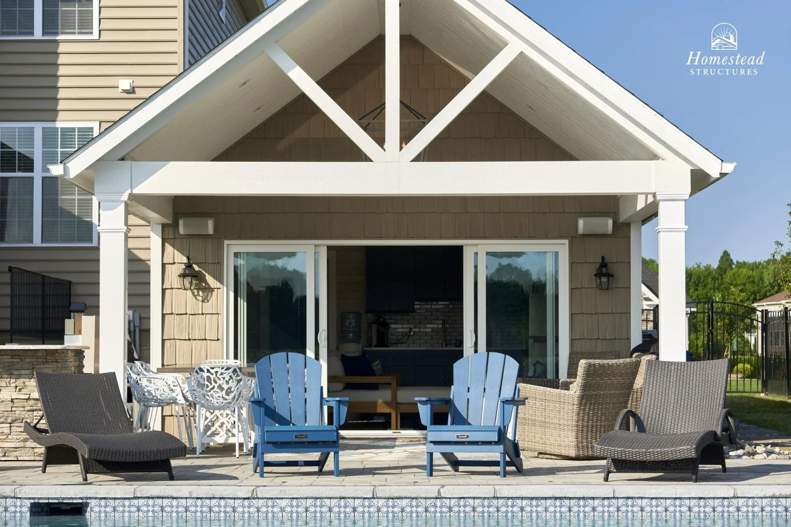 Back patio of a house with outdoor furniture including Adirondack chairs and lounge chairs, sliding glass doors, and a visible pool in the foreground
