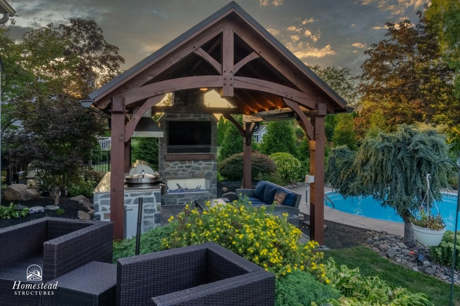 Outdoor backyard with a wooden gazebo, seating furniture, a built-in grill and a fireplace under the gazebo, a swimming pool, and lush greenery during sunset.