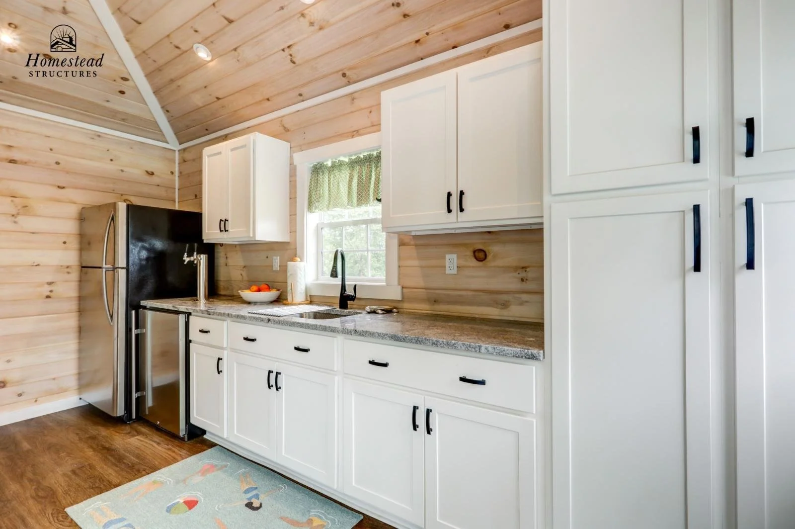 Kitchen with wood-paneled ceiling, white cabinets with black handles, granite countertop, stainless steel refrigerator, window with green curtain, black faucet, bowl of fruit, paper towels, and light-colored rug on hardwood floor.