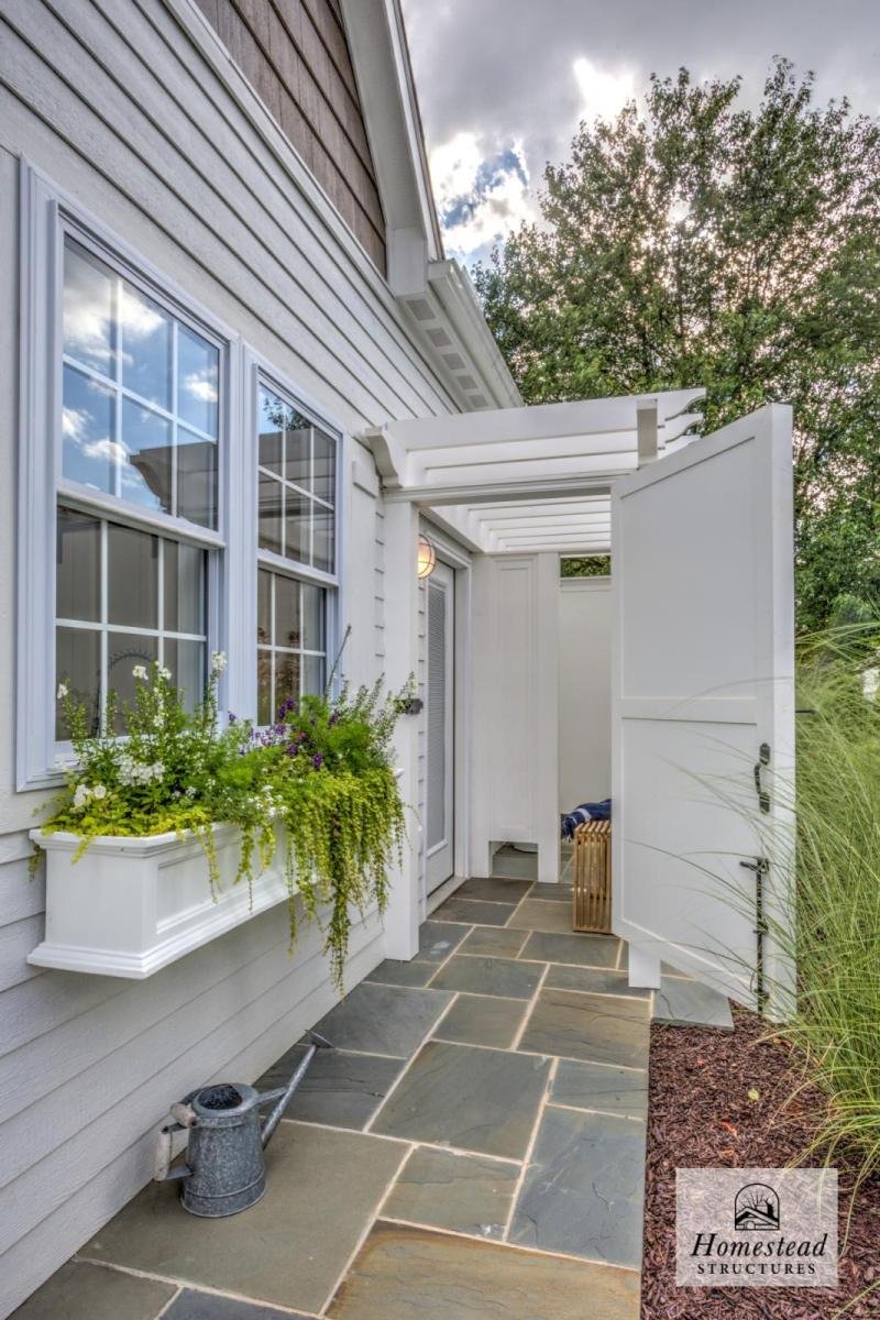 A cozy outdoor porch area with a white pergola, potted flowers in a window box, a sliding screen door, and a stone pathway leading to a garden with tall grasses and trees.