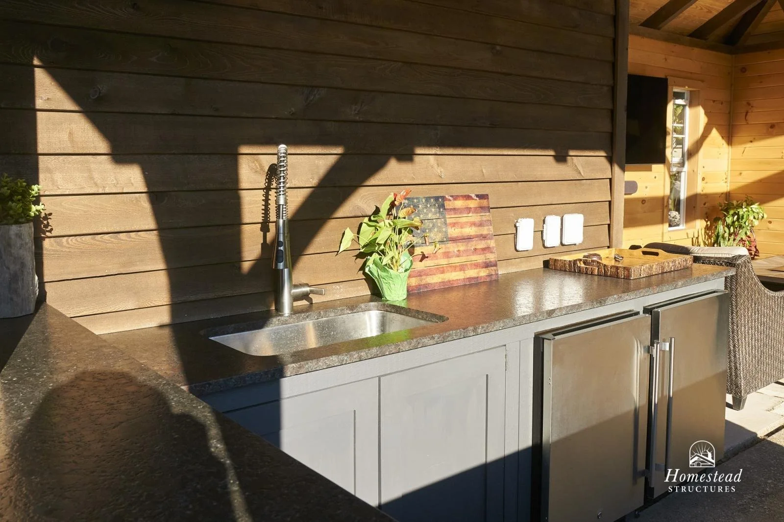 Outdoor kitchen with gray cabinets, a granite countertop, a stainless steel sink, a modern faucet, a small potted plant, an American flag design wooden cutting board, three white electrical outlets, a wooden tray, and a wicker chair. Wooden wall pane