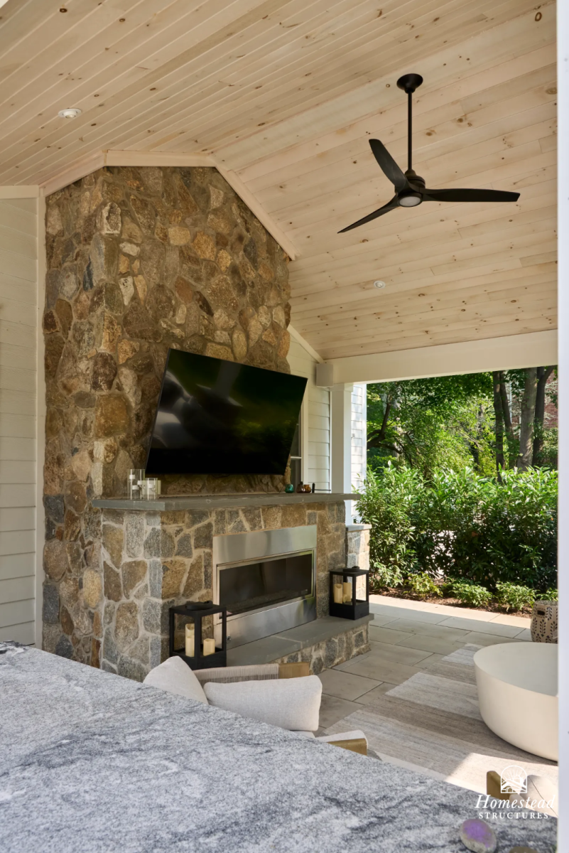 Covered outdoor living space with a stone fireplace and a mounted flat-screen TV, ceramic lanterns with candles, a white freestanding bathtub, ceiling fan, and lush green trees outside.