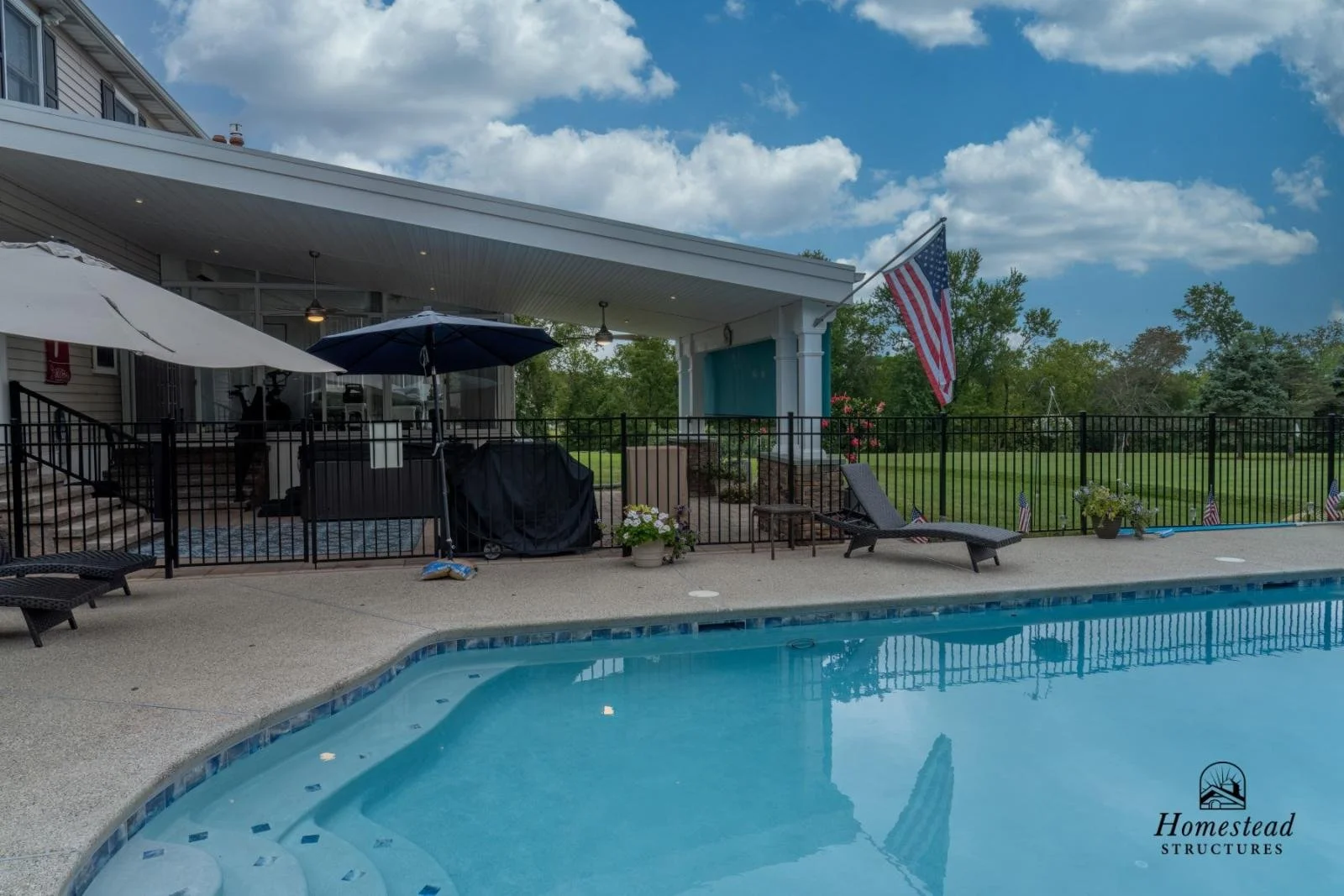 A backyard swimming pool scene with a black metal fence, lounge chairs, and potted plants, a house with a covered patio, and an American flag; cloudy sky and green trees in the background.