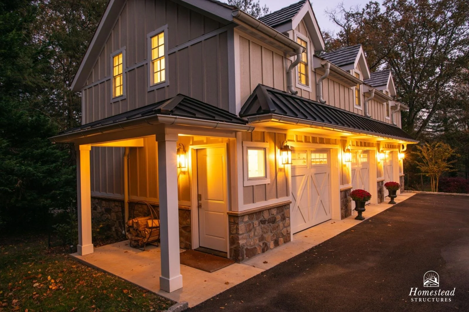 A modern two-story garage with a barn-style design, gray vertical siding, stone foundation, and white trim, illuminated by warm outdoor lighting at dusk, with potted plants near the doors and driveway visible in the foreground.
